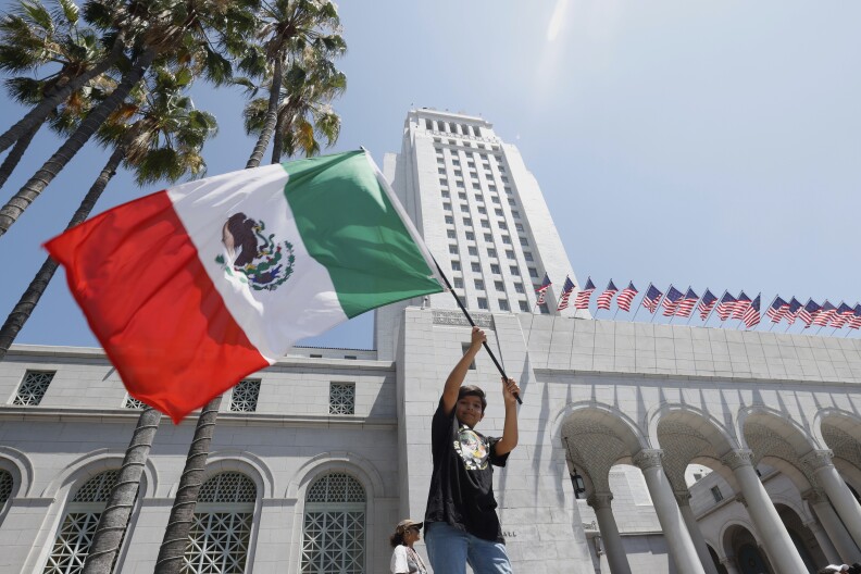A boy in a black T-shirt and jeans waves the Mexican flag in front of a stone building. American flags line the building behind him.