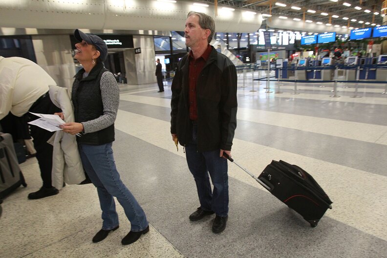 Two men at an airport, one of them holding papers in his hand looking up at something off camera, while another behind him holds a rolling suitcase.