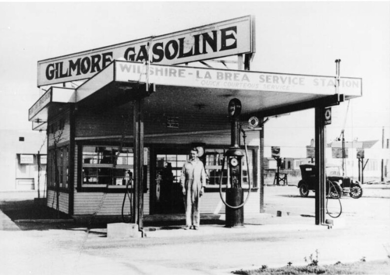 The station's office is covered by clapboard veneers, and signs on the edges of its roof's overhang reads, "Wilshire-La Brea Service Station. Quick Courteous Service." A sign on the top of the building reads "Gilmore Gasoline." A parked automobile is visible in the right background.