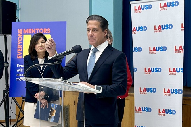 Superintendent Alberto Carvalho stands at a clear lectern with a microphone in front of a portable screen with the letters "LAUSD" printed in red and blue. He wears a navy suit, a light blue tie, and a white collared shirt. His hair is neatly slicked to one side. A woman in a white skirt and a dark blazer stands off to one side with her hands folded in front of her, and she's holding a brochure. Both have a light or olive complexion.