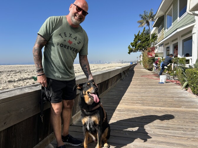 A male presenting person holds a black and brown dog. They stand on a wood boardwalk.