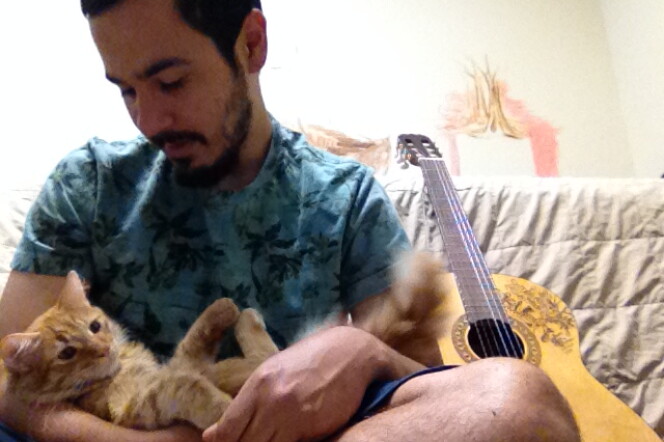 Frank Campos is seated holding a cat. A guitar sits to his left. 