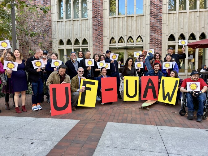 A large group of people with diverse skin tones are arranged in two rows in front of a brick building. The back row hold up signs with a union logo, while the front row hold up large pieces of paper that spell out "UF-UAW."