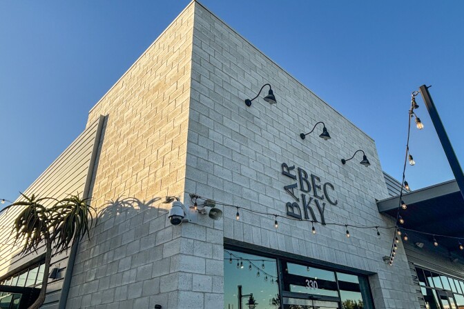 The exterior of Bar Becky, a modern gray brick building with black lamps, string lights, and a clear blue sky above.