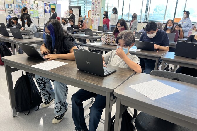 A classroom full of teenagers sit at long tables and stare at laptop screens in a classroom.