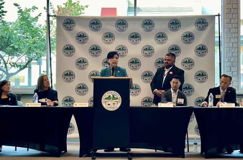 An Asian American woman in an aqua suit stands at a podium with two people seated on both sides of her and one man standing to her left.