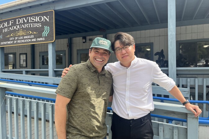 Two Asian men, on in a ball cap, embrace side-by-side and smile for a photo.