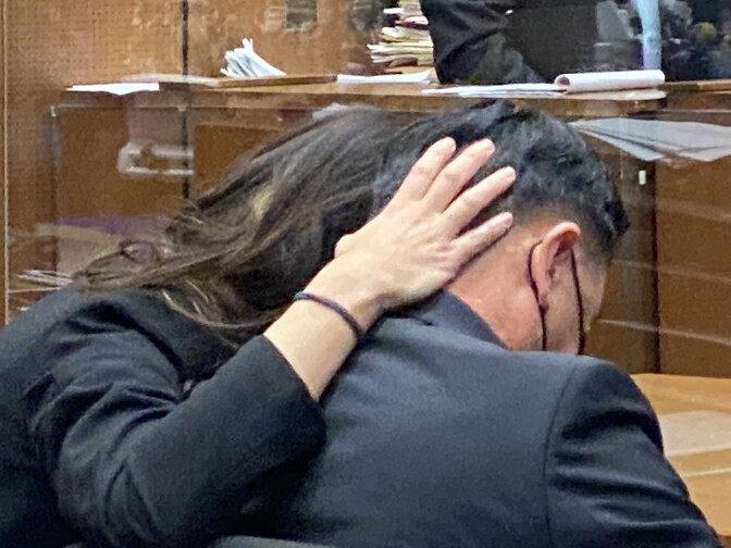 A woman has her hand on the head of a man wearing a black mask. Both are seen from the back and wearing dark suits.