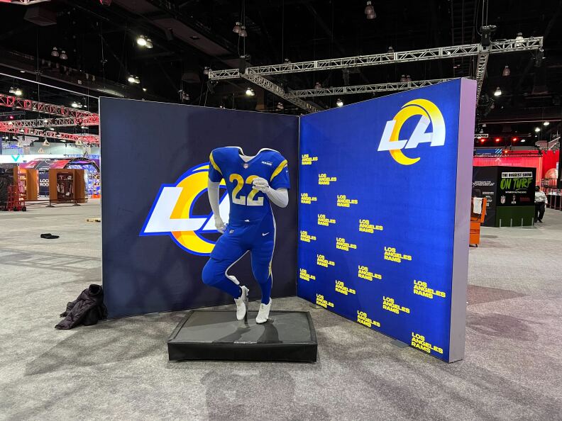 A cardboard cutout of a blue Los Angeles Rams jersey with the head space cut out so visitors can stand behind it and appear to be wearing the uniform is placed on a raised display in front of screens with the Rams logos at the NFL Super Bowl Experience.