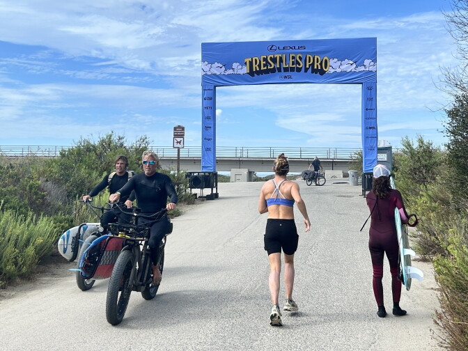 People walk and ride bikes with surfboards near a beach path arch reading 'Trestles Pro' for a surfing competition.