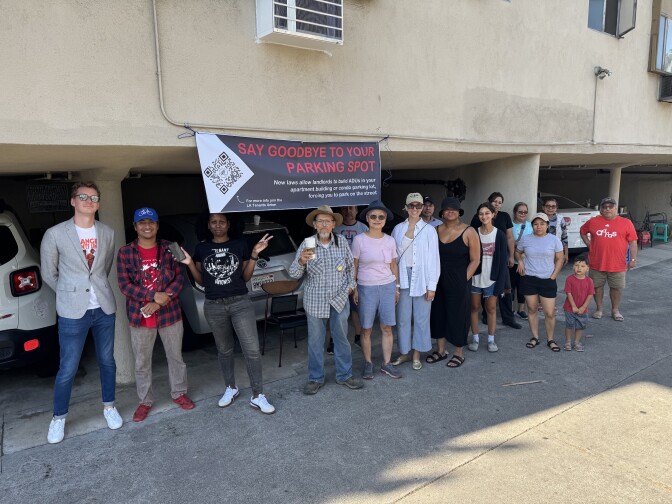 A group of people stand in front of low-level parking lots under an beige apartment complex. A sign hangs above one parkign spot that says, "Say goodbye to your parking spot." 