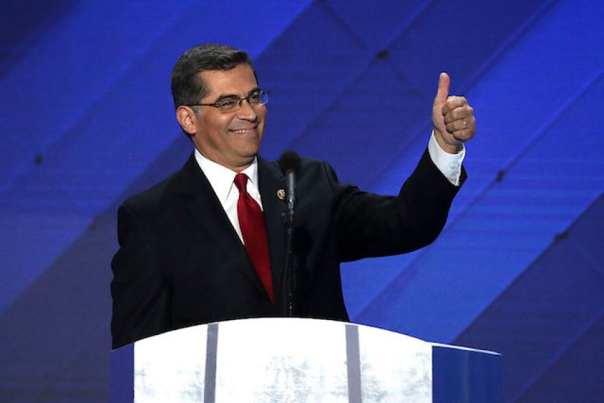 A man wearing a black suit jacket,red tie and white shirt stands at a podium, smiling while holding his left thumb up in the air
