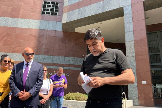 A Latino man in a black T-shirt and pants opens a piece of papaer while other look on outside a building.