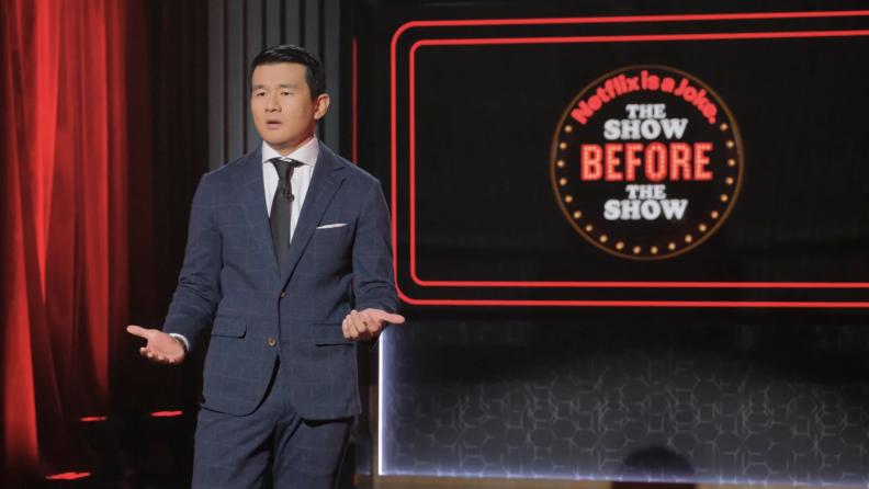 A man with lighter skin tone and a grayish-blue suit stands on a stage in front of an illuminated sign that reads "Netflix is a Joke: The Show Before The Show."