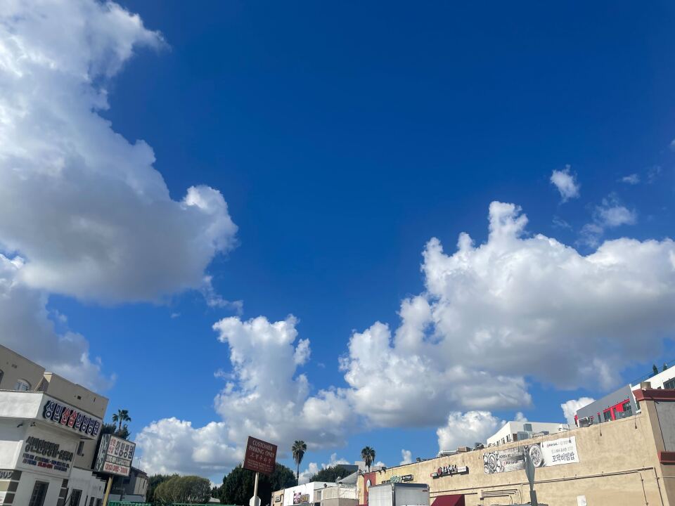 Clouds flank a cobalt blue midday sky, with signs in English and Korean on a strip mall in the bottom part of the image.