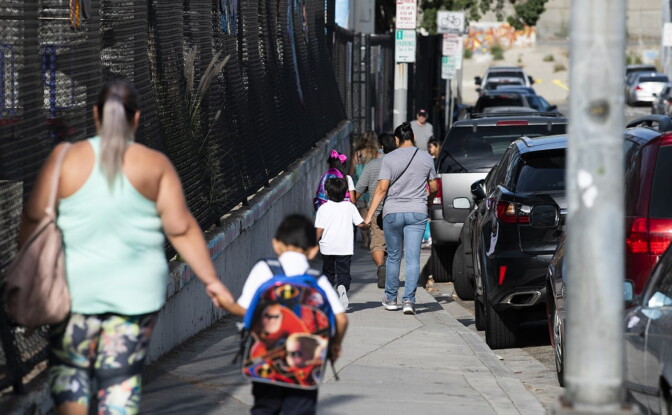 parents walk their children along a sidewalk with a chainlink fence on one side and a row of cars on the other side.