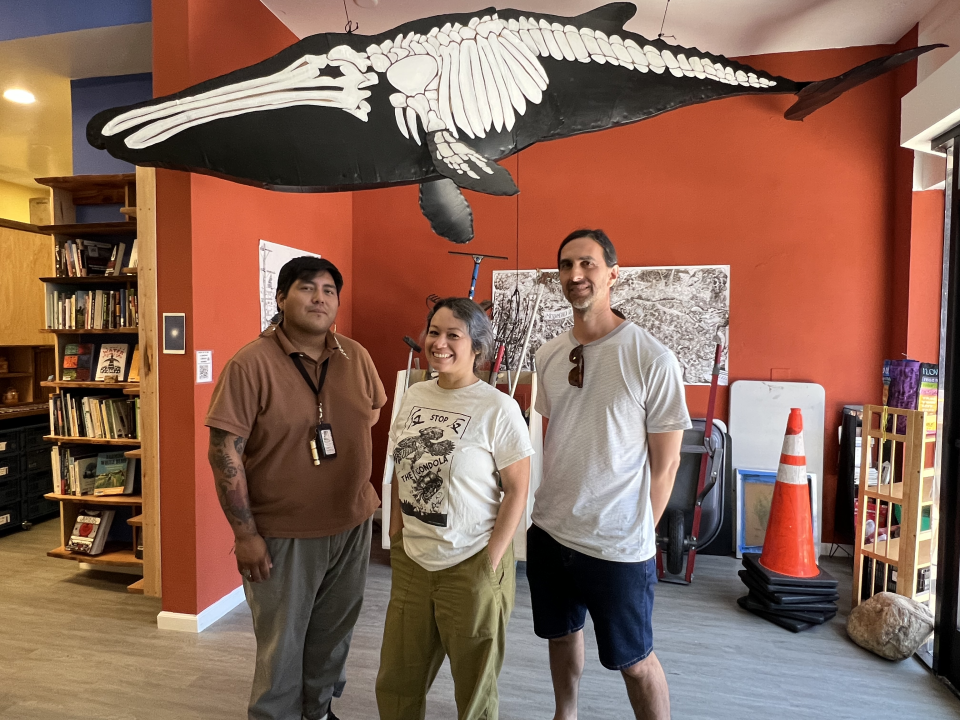 Lending Library Manager César Montero, Executive Director of Coyotl+Macehualli Brenda Contreras, and co-founder of Coyotl+Macehualli Micah Haserjian stand below a recreation of an ancient whale found in Northeast L.A. by the artist Sandra de la Loza.
