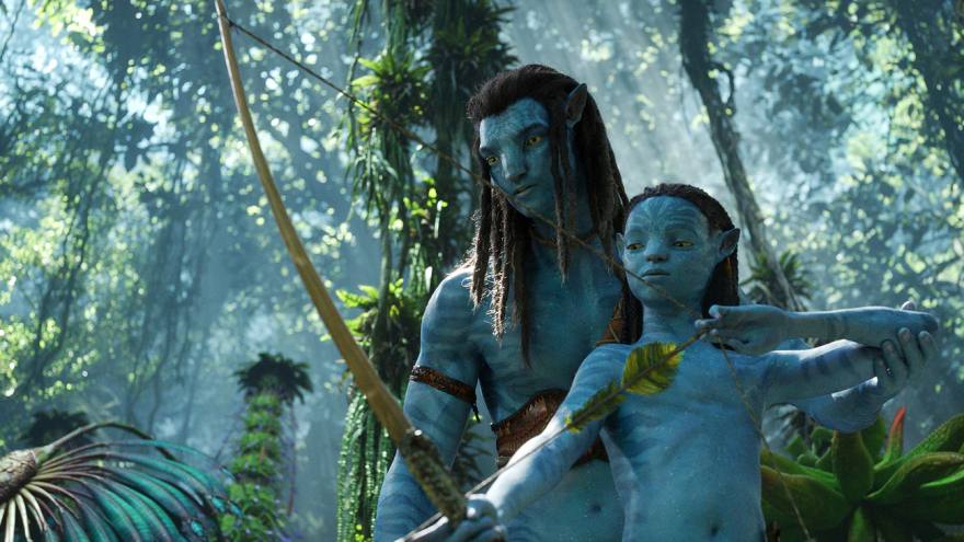 Sam Worthington and Jamie Flatters stand in a green, tropical forest with tall trees and lush plants. They both have blue skin, pointed ears, and long black hair, signature features of the Na'vi, the native inhabitants of the planet Pandora. Worthington stands behind the shorter and younger Flatters. Flatters holds a bow in his right hand with the strong of the bow and an arrow with leaf feathers in his left. His right arm is extended towards the ground and his left arm is bent as he draws back the arrow, preparing to fire. Worthington holds Flatters' left elbow up, guiding him, teaching him how to aim and shoot. They both look toward the ground where the arrow is aimed. 