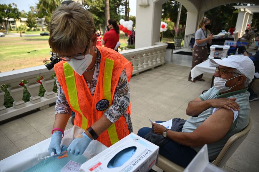 On the left, a nurse wearing an orange safety vest, a mask, and gloves prepares a syringe of COVID-19 vaccine. On the right, a person wearing a mask pulls up his sleeve in preparation for the shot. They are outdoors on a porch -- in the background, there are other similar vaccination stations and beyond that is a lawn.