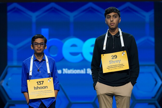 Two teens stand next to each other. The one on the right is much taller. Each has their first name and a number on a lanyard around their necks.