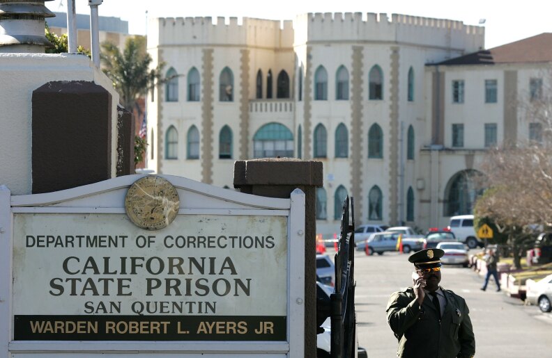 A man wearing a guard uniform holds a phone to his ear next to a sign that says: California State Prison San Quentin.