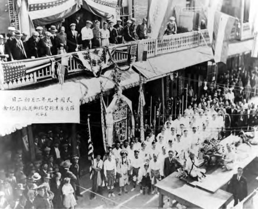 In an old black and white photo, a dance troupe dressed in white shirts makes a formation of three close lines in the middle of a street in Old Chinatown. People look on from balconies over the street.