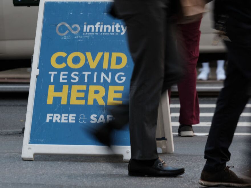 A sign reads COVID TESTING HERE as pedestrians pass by