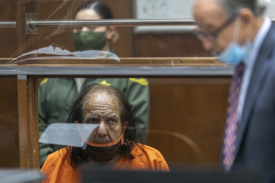 A white man who appears to be in his 60s or 70s sits behind a plastic partition in a court room. He wears an orange prison jumpsuit and black face mask. His hair is black and long on the sides, balding on top. He looks concerned and confused. A man wearing a suit can be seen blurred in the foreground.