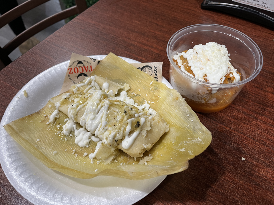 A tamale sits on a paper plate with Mexican crema drizzled over it, on a table