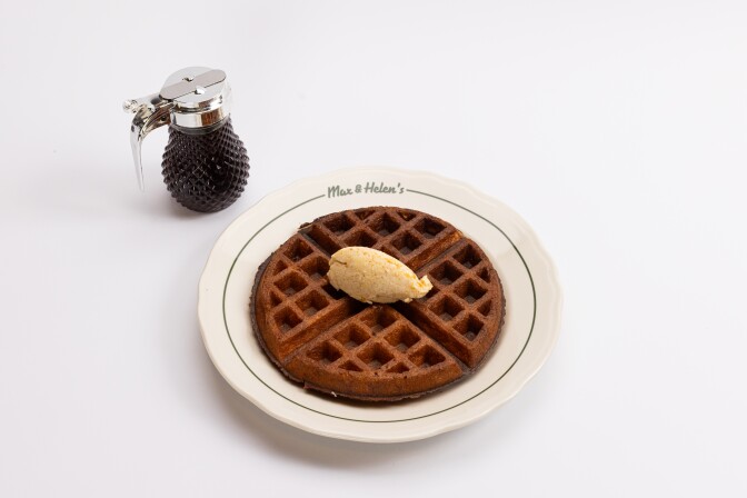 A golden-brown waffle on a white plate with 'Max & Helen's' printed on the rim, topped with a quenelle of whipped maple butter, with a small pitcher of syrup beside it.