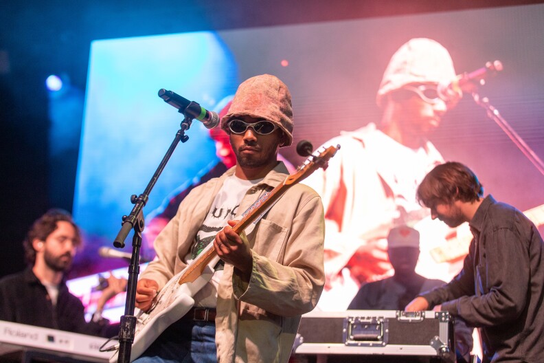 Singer Toro y Moi plays a white guitar on stage with his band. He's wearing a tan jacket, pink bucket hat, and sunglasses. The rest of the band's members are dressed in black.