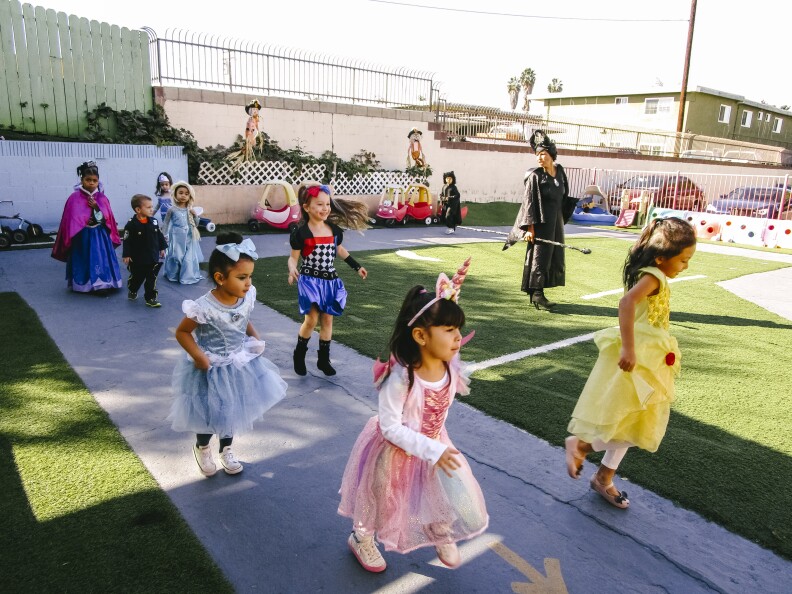 In this image, a group of children dressed in costumes walk together outside with a teacher.