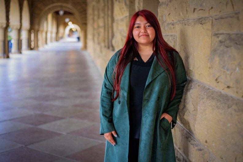 A young woman with Brown skin and long, dyed red hair stands in a stone walkway that is covered with arches held up by columns. She wears a long turquoise jacket and a black shirt.