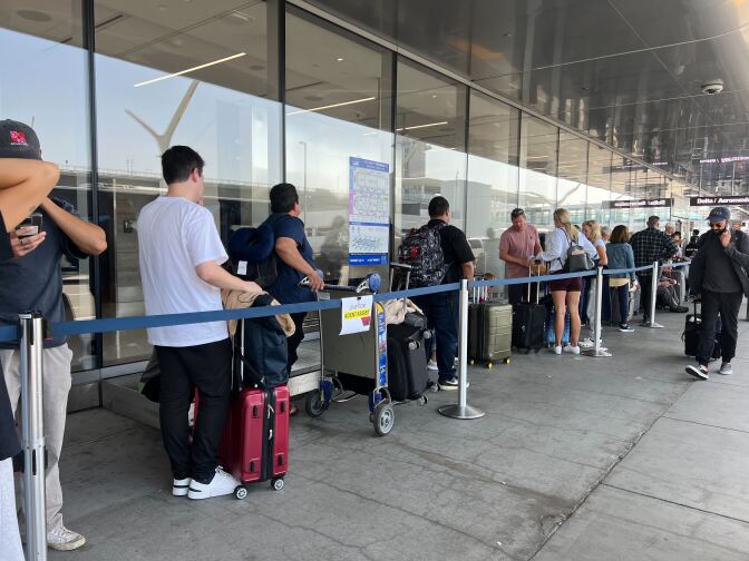 Passengers holding on to wheeled suitcases stand in a line behind blue tape. Some passengers carry backpacks. 