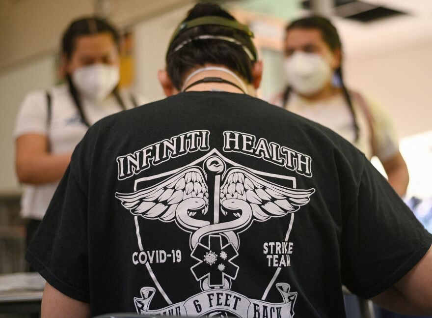 A person's back is to the camera wearing a black T-shirt that reads: Infiniti Health COVID-19 Strike Team