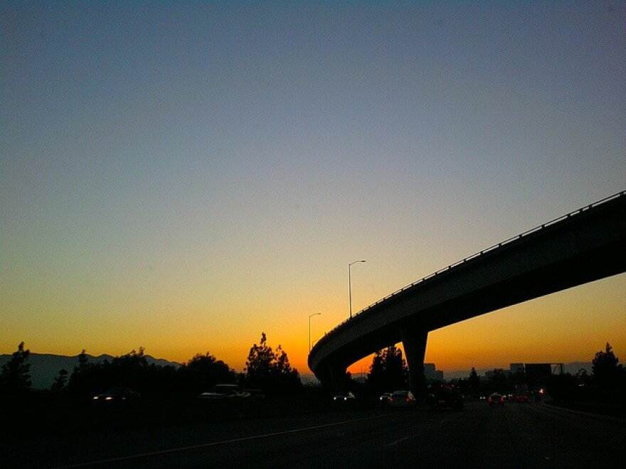 The sunset turns the sky orange in the background. In the foreground is the silhouette of a freeway bridge.