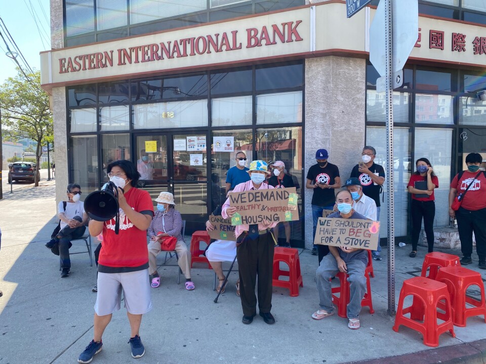 Kris Chan, one of the protest organizers, is wearing a face mask, red shirt and gray shorts while holding a megaphone in front of Eastern International Bank. Other tenants and organizers are standing and seated behind Kris while holding sings that say "we demand healthy and habitable housing" and "We shouldn't have to beg for a stove."