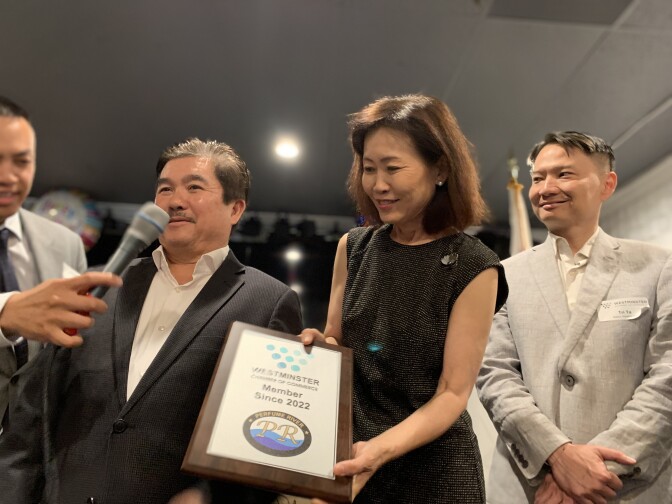 An Asian American woman in her 60s wearing a black shift dress presents a plaque from the Westminster Chamber of Commerce to an Asian American man in a suit.