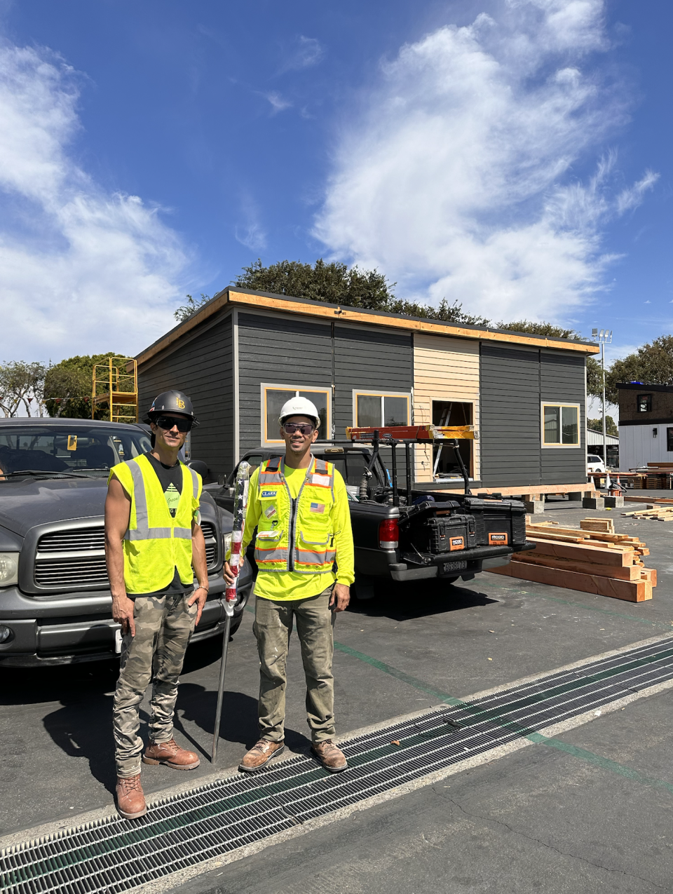 Workers representing the California State University, Long Beach team stand in front of two trucks in front of their "sustainable home". Both are wearing camo pants, yellow reflective safety vests, sunglasses and hard hats. The one on the left has a bronze hard hat while the one on the left has a white hard hat. The home behind them looks nearly complete and is a a gray sloped building with a sandy colored trim along the roof. There is wood scattered directly in front of the house.