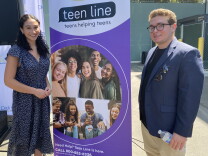 Teen Line counselors Lily Kramon (L) and Jonathan Gelfond (R) completed 65 hours of training and months of shadowing in order to support their peers over calls and texts. They stand on either side of a banner that features pictures of youth hanging out and smiling. The banner reads "teen line, teens helping teens." 