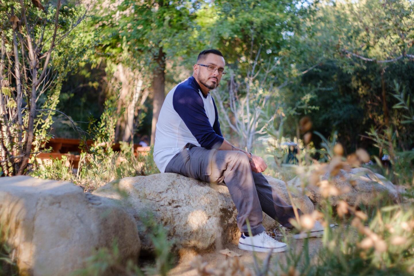 José Norrington, wearing glasses, white sneakers, khaki pants and a white t-shirt with long blue sleeves, sits on a boulder looking at the camera at L.A.'s Vista Hermosa Natural Park. There is another boulder in the foreground, and trees, grass and shrubbery in the background.