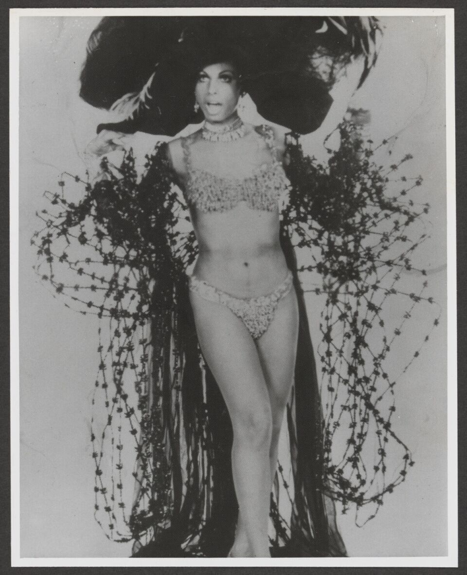 A black and white publicity shot of trans woman performer Sir Lady Java in a bejeweled bikini and intricate cape.