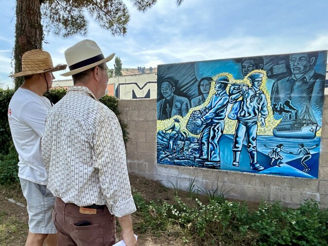 Two people wearing hats stand in front of a mural painted in blue.