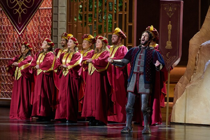 A group of singers in two rows on a stage, with arms crossed in front of them and wearing red robes and hats, both with gold accents. To their left is singer-songwriter Josh Groban in a medeival style outfit with partial knight armor, with his arms outstretched and mouth open in song.