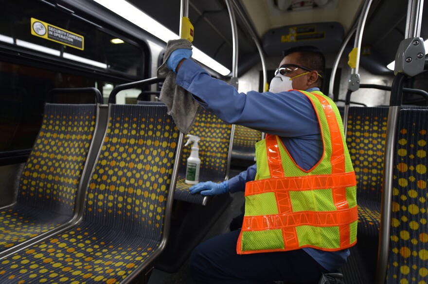 A Metro worker wipes down a metal pole inside a subway train. He wears a neon orange and yellow vest, a face mask and blue latex gloves.