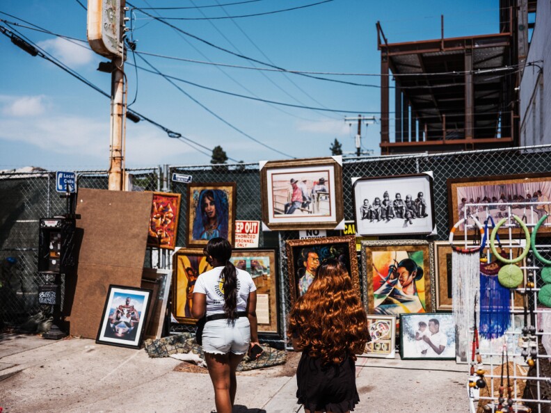 Two Black women, one with a pony tail and the other with wavy red hair, approach a chainlink fence that has a series of paintings and other framed artwork on display.
