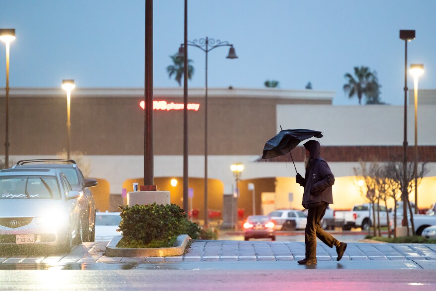A man wearing black pants and black jackets, holds a black umbrella while walking down a side walk in front of a parking lot with a CVS Pharmacy.