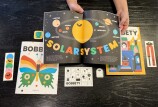A pair of hands with medium light skin tone hold open a brightly colored magazine depicting the solar system with a bright yellow circle surrounded by smaller circles in different colors. The magazines in the background have a butterfly and ghosts on the cover. 