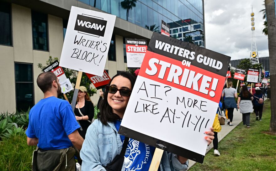 A light skinned woman wearing sunglasses and a denim shirt smiles at the camera as she holds a sign that says WRITERS GUILD ON STRIKE. AI? MORE LIKE AY-YI-YI! Other picketers stand behind her, in front of a big office block with reflective glass windows