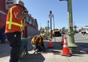 There are two men in high-visibility vests and wide-brimmed hats on a road. One is standing next to a blue wheelbarrow with a shovel. The other man is on his knees using a tool to spread wet concrete over box where street lighting wires are stored. 